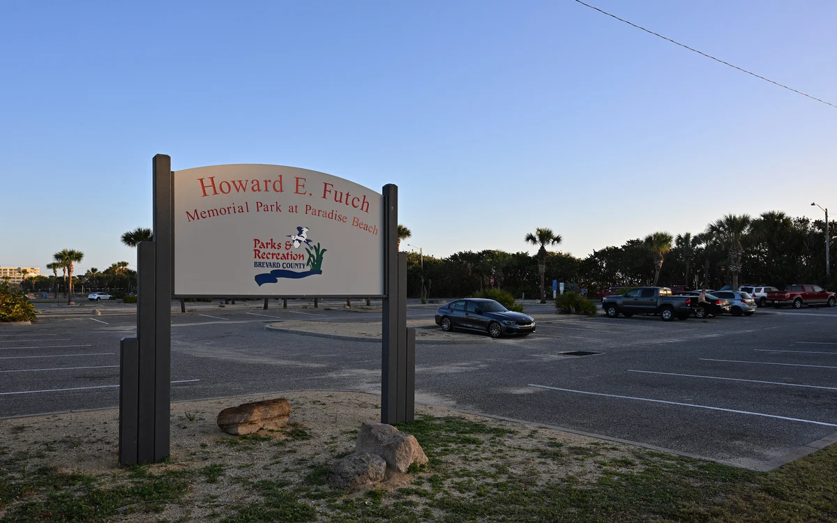 The Brevard County Parks entrance sign for Howard E. Futch Memorial Park at Paradise Beach, with the parking lot, palms and sea grape behind it