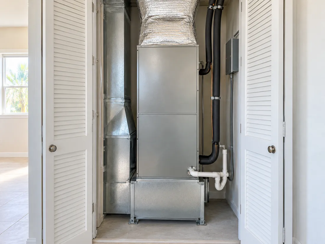 An air handler standing in an open hall closet with insulated ducting above it and a drain line at its foot