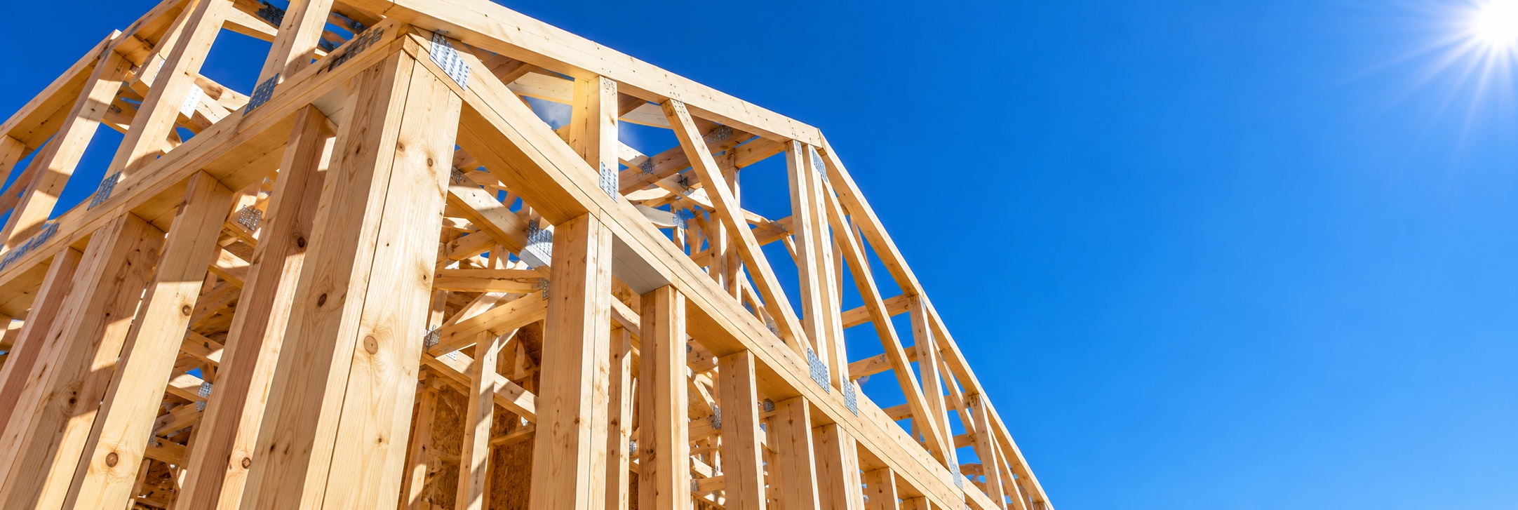 Timber roof framing of a house under construction, seen from below against a clear blue sky